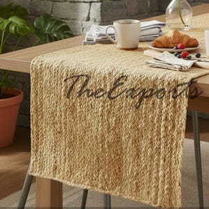 May include: A dining table set with a woven beige table runner. The table is set with two white mugs, a croissant, and a French press. A vase with white flowers sits on the table. The text "The Exports" is on the table runner.