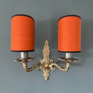 Op de afbeelding: Een paar gouden wandlampen met oranje stoffen lampenkappen. De lampen hebben een decoratief scroll-ontwerp en zijn aan een muur gemonteerd.
