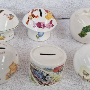 Puede incluir: Seis alcancías de cerámica con diferentes diseños. Los diseños incluyen un hongo con personajes de Winnie the Pooh, una oruga en una manzana, una alcancía redonda con la inscripción 'It's a lovely day' y una alcancía redonda con un diseño de Thomas the Tank Engine.