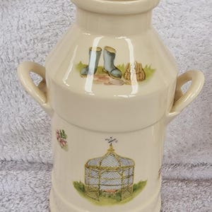 May include: A white ceramic vase shaped like a milk jug with two handles. The vase has a floral design with a birdcage, boots, and a basket.