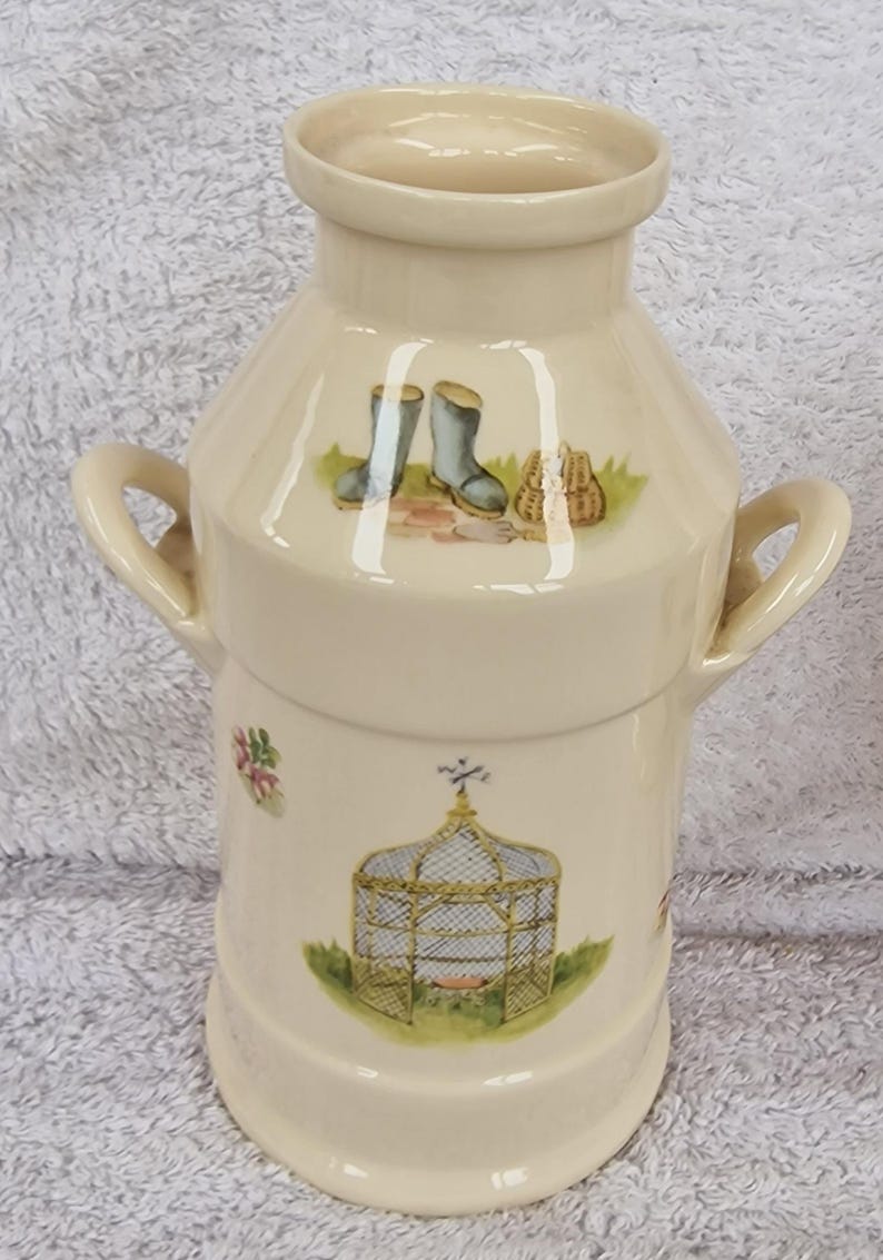 May include: A white ceramic vase shaped like a milk jug with two handles. The vase has a floral design with a birdcage, boots, and a basket.
