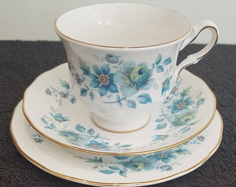 May include: A white teacup, saucer, and dessert plate set. The set features a floral pattern in shades of blue and green with gold trim. The teacup has a curved handle and sits atop a pedestal base.