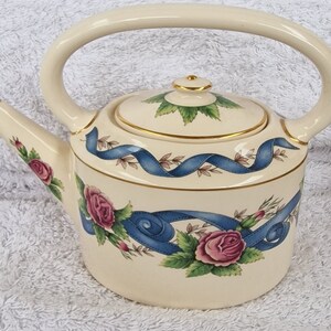 May include: A white ceramic teapot with a blue ribbon and rose design. The teapot has a lid with a gold trim and a handle.