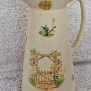 May include: A white ceramic pitcher with a garden theme. The pitcher has a handle and is decorated with a green watering can, a brown basket, a wooden rake, a wooden gate, and pink and yellow flowers.