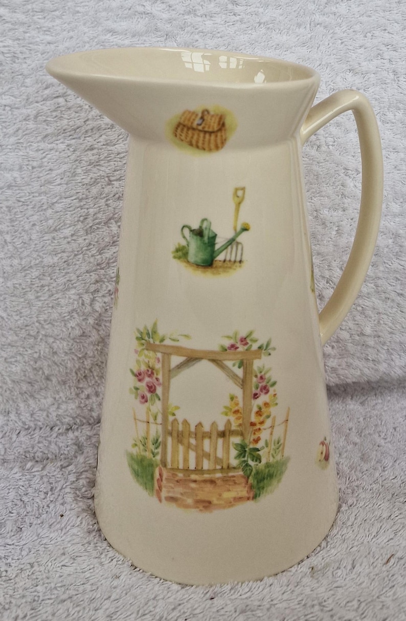 May include: A white ceramic pitcher with a garden theme. The pitcher has a handle and is decorated with a green watering can, a brown basket, a wooden rake, a wooden gate, and pink and yellow flowers.