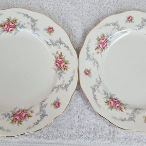 Könnte beinhalten: Zwei weiße Porzellanteller mit gewellten Rändern und Goldrand. Jeder Teller hat ein Blumendesign mit rosa Rosen, blauen Akzenten und einem grauen Rankenmuster am Rand. Die Teller sind Teil eines Geschirrsets.