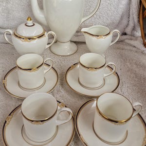 May include: A white porcelain tea set with gold trim and navy blue detailing. The set comprises a teapot, sugar bowl, creamer, and four cups with saucers. The tea set is displayed on a white surface.