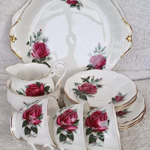 Pode incluir: Um conjunto de chá de porcelana branca vintage com um padrão floral de rosas vermelhas. O conjunto inclui um prato de servir grande, um cremeira, xícaras e pires, todos com detalhes dourados. As rosas são de um vermelho profundo.