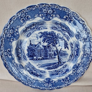 May include: A blue and white ceramic plate with a detailed floral border. The centre of the plate features a blue and white illustration of a house with a tree and horses in a field. The text "The Precinct" is visible in the centre of the plate.