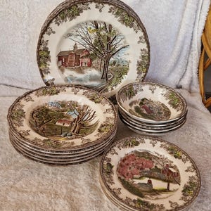 Puede incluir: Un conjunto de platos y cuencos de cerámica vintage. Los platos presentan un diseño de paisaje escénico con un granero rojo, árboles y un camino sinuoso. Los bordes están decorados con un patrón floral marrón. El juego incluye platos llanos, platos de ensalada y cuencos pequeños.