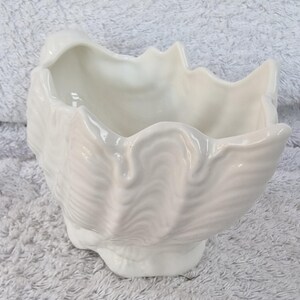 May include: A white ceramic planter shaped like a seashell with a ruffled edge. The planter is designed to look like a clam shell.