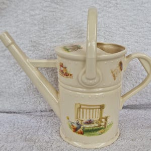 May include: A white ceramic watering can with a handle and a spout. The can has a floral design with a bench, a wheelbarrow, and other gardening tools.