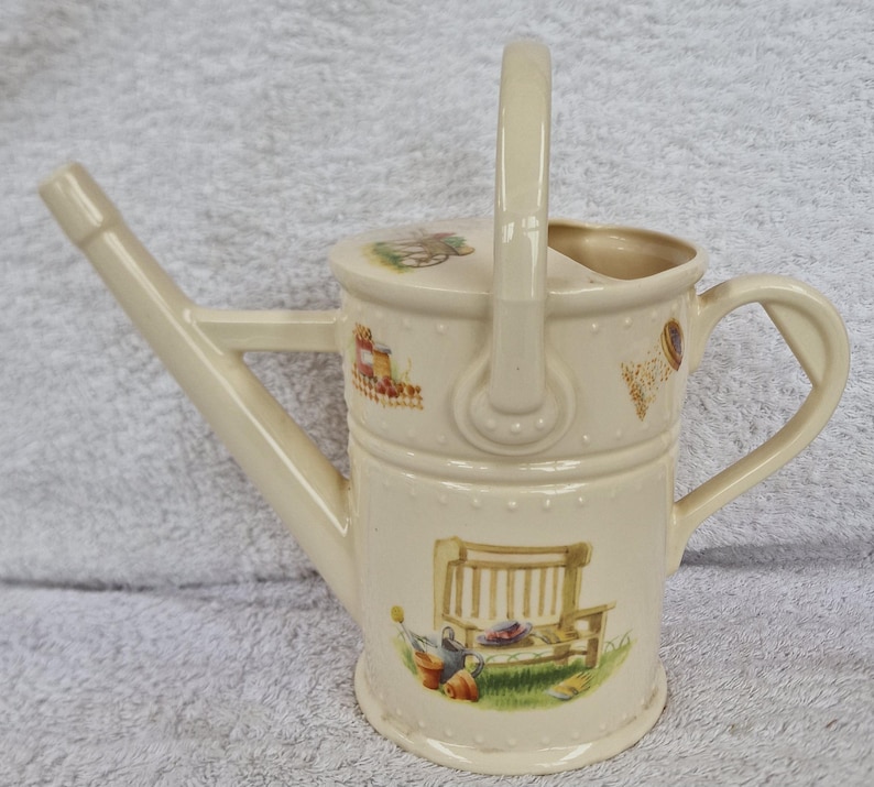 May include: A white ceramic watering can with a handle and a spout. The can has a floral design with a bench, a wheelbarrow, and other gardening tools.