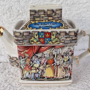Może przedstawiać: Biały ceramiczny czajnik w kształcie zamku z czerwono-złotym wzorem. Czajnik przedstawia scenę z dworu króla Artura w Camelocie, z postaciami takimi jak król Artur, królowa Ginewra i Sir Lancelot. Czajnik ma złoty uchwyt i dzióbek.