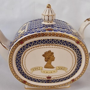 Può includere: Teiera color crema con un motivo geometrico blu e oro. Il centro ovale presenta un profilo dorato di una figura incoronata e le date 1952 e 2002. La teiera ha un beccuccio, un manico e un coperchio rifiniti in oro.