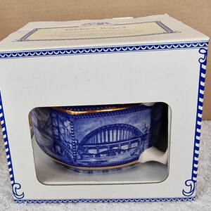 Puede incluir: Una tetera de cerámica azul y blanca con un diseño de puente, presentada en su caja original. La tetera tiene ribetes dorados y una detallada ilustración de un puente. La caja es blanca con detalles azules y el texto "Bridges Teapot".
