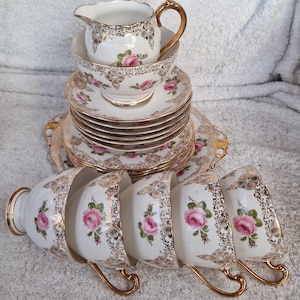 Può includere: Un servizio da tè vintage con tazze, piattini e una lattiera in porcellana bianca. Ogni pezzo è decorato con rose rosa e dettagli in filigrana dorata. Il set include più tazze, piattini e una lattiera, tutti impilati.
