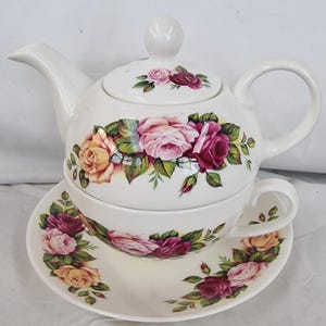 Pode incluir: Um conjunto de chá em cerâmica branca para uma pessoa, composto por um bule que se empilha sobre uma chávena, que repousa num pires a condizer. O conjunto apresenta um padrão floral de rosas cor-de-rosa, amarelas e vermelho-escuras.