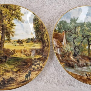 Puede incluir: Dos platos decorativos con borde dorado que presentan pinturas de paisajes. El primer plato representa una escena rural con árboles, un campo y un grupo de personas. El segundo plato muestra una cabaña junto a un río con árboles y un cielo nublado.