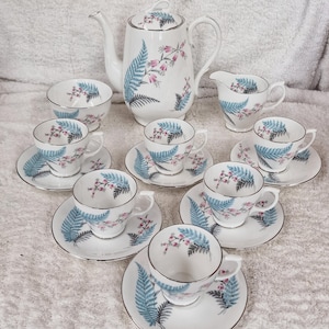Puede incluir: Un juego de té vintage de porcelana blanca con un diseño floral. El juego incluye una tetera, una jarra de crema, un azucarero y varias tazas y platillos. El diseño presenta hojas de helecho azules y flores rosas. Los bordes son plateados.