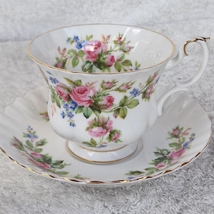 Könnte beinhalten: Weißes Tee-Set mit Tasse und Untertasse, verziert mit rosa, blauen und grünen Rosenmustern. Die Tasse hat einen goldenen Rand und Henkel, die Untertasse einen gewellten Rand und einen goldenen Zierrand. Ein klassisches Teeservice.