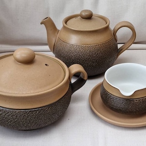 Puede incluir: Una tetera de cerámica marrón y beige, una cacerola de cerámica marrón y beige con tapa y una salsera de cerámica marrón y beige con interior blanco y un platillo.