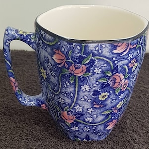 Puede incluir: Una taza de cerámica con un fondo azul denso de textura punteada, decorada con un patrón repetitivo de rosas rosadas, pequeñas flores blancas y flores amarillas. La taza tiene un interior de color crema y un borde oscuro en la parte superior.
