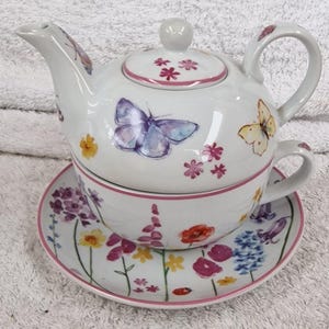 May include: A white ceramic tea-for-one set with a teapot, teacup, and saucer. The set is decorated with watercolour-style butterflies and floral designs in purple, yellow, and pink. The set includes a lid and a handle.