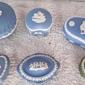 Könnte beinhalten: Sammlung blauer und weißer Keramik-Schmuckdosen in verschiedenen Formen, darunter rund, oval und herzförmig. Jede Dose hat ein erhabenes weißes Reliefdesign und einen dekorativen weißen Rand. Eine grüne Dose ist ebenfalls enthalten.