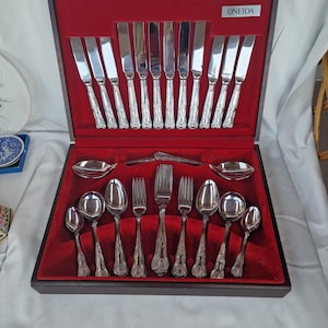 Könnte beinhalten: Ein Set aus silberfarbenem Edelstahlbesteck in einem dunklen Holzkasten mit rotem Samtinnenfutter. Das Set enthält Messer, Gabeln und Löffel mit verzierten Griffen. Ein kleines Schild mit der Aufschrift ONEIDA ist deutlich zu erkennen.