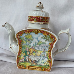 Può includere: Una teiera in porcellana bianca con un design floreale rosa e oro. La teiera presenta una scena di persone che prendono il tè pomeridiano in un giardino. La teiera ha un coperchio con un pomo dorato e le parole "Afternoon Tea" stampate sul fondo.