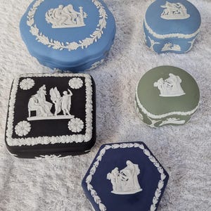 May include: A selection of decorative trinket boxes in various shapes and colours. The boxes are blue, black, and green, with white relief designs and floral borders. The shapes include round, square, and hexagonal.