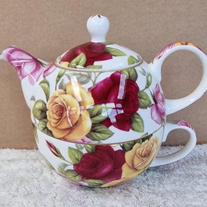 May include: A white porcelain tea set featuring a teapot and teacup, both adorned with a floral rose pattern in shades of yellow, red, and pink. The teapot has a curved spout and handle, with a matching lid and a small knob. The teacup is stacked below the teapot.