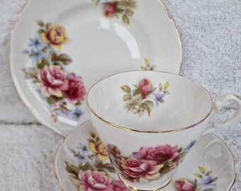 May include: A white teacup and saucer set with a floral pattern featuring pink, yellow, and blue roses. The set has a gold trim around the edges.