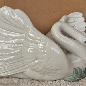 May include: A white ceramic swan figurine with detailed feather textures. The swan is in a resting pose, with its neck curved and head tucked. The figurine has a glossy finish and is accented with subtle gray and green details.