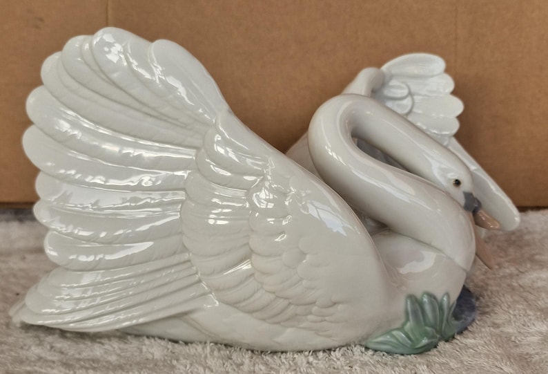 May include: A white ceramic swan figurine with detailed feather textures. The swan is in a resting pose, with its neck curved and head tucked. The figurine has a glossy finish and is accented with subtle gray and green details.