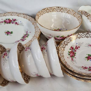May include: A collection of white porcelain tea service pieces featuring pink rose floral patterns and ornate gold filigree trim. The set includes multiple saucers, tea cups, a sugar bowl, and a creamer pitcher with a handle, all with scalloped edges.