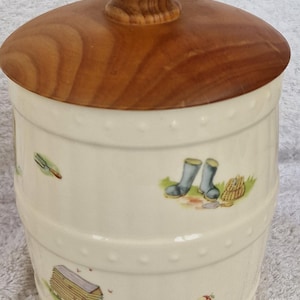 May include: A white ceramic jar with a wooden lid. The jar has a country-themed design with images of boots, a rake, a barn, and an apple.