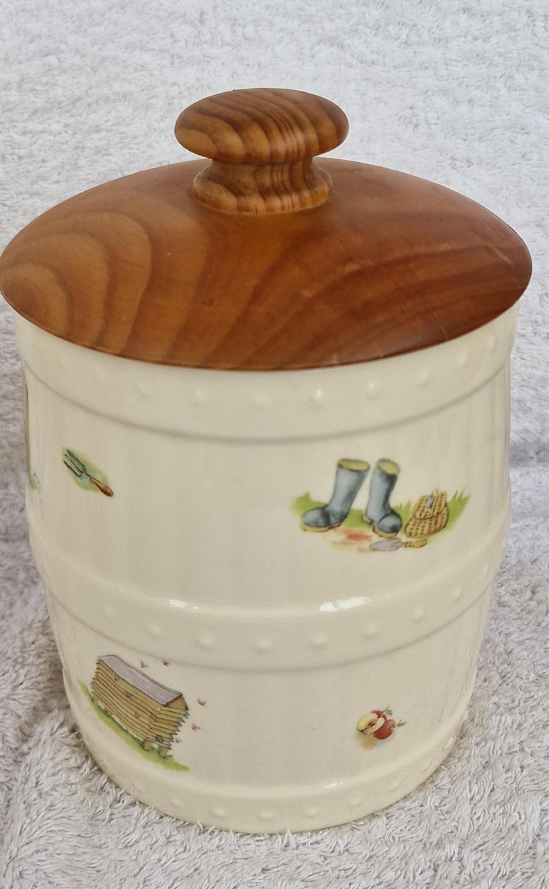May include: A white ceramic jar with a wooden lid. The jar has a country-themed design with images of boots, a rake, a barn, and an apple.