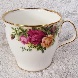 Puede incluir: Taza de té blanca con borde y asa dorados, adornada con un diseño floral de rosas rosas y amarillas. La taza tiene una forma clásica, perfecta para disfrutar del té o café.
