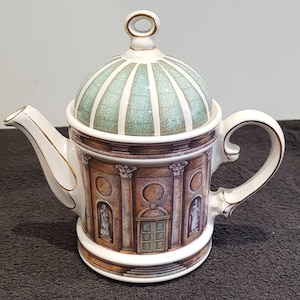 May include: A ceramic teapot featuring a domed lid with a green and white crackle glaze pattern. The body displays a detailed architectural illustration of a classical building with columns, arched doorways, and statues, accented with gold trim.