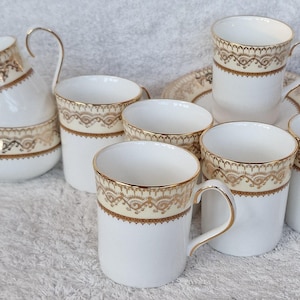 Puede incluir: Un juego de té de porcelana blanca con detalles de filigrana dorada. El juego incluye una lechera, seis tazas y un platillo. Las tazas tienen un asa y una banda decorativa alrededor del borde. La lechera tiene un asa curva.