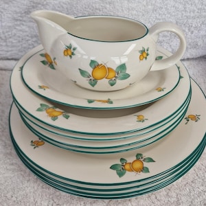 Pode incluir: Um conjunto de seis pratos de cerâmica branca com uma borda verde e um design de limão. Os pratos estão empilhados uns sobre os outros, com um molho a condizer no topo.