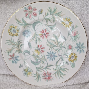 Può includere: Piatto rotondo in ceramica bianca con bordo dorato, decorato con un motivo floreale dai colori pastello. Il design presenta fiori rosa, gialli e blu con foglie e steli verdi. Piatto decorativo in stile vintage.