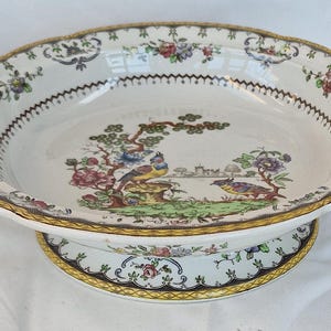 May include: A white ceramic serving dish with a pedestal base. The dish features a colourful bird and floral design with a gold rim. The rim has a scalloped edge and decorative accents.