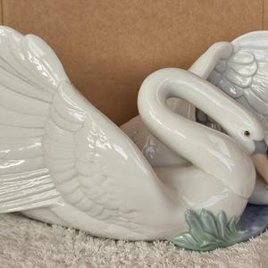 May include: A white ceramic swan figurine with outstretched wings and a curved neck. The swan has a detailed feather design and a light blue base with green accents. The beak is a light tan color.