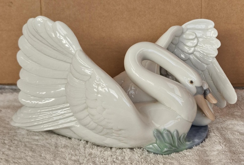 May include: A white ceramic swan figurine with outstretched wings and a curved neck. The swan has a detailed feather design and a light blue base with green accents. The beak is a light tan color.