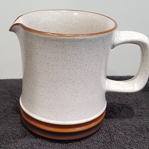 Puede incluir: Una jarra de cerámica blanca con un esmalte moteado marrón y un borde marrón. La jarra tiene un asa y una banda marrón alrededor de la base.