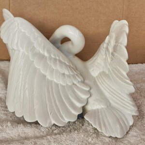May include: A white ceramic swan figurine with outstretched wings. The swan's wings are detailed with individual feather carvings. The swan's neck is curved, and the head is turned back. The figurine is a decorative item.