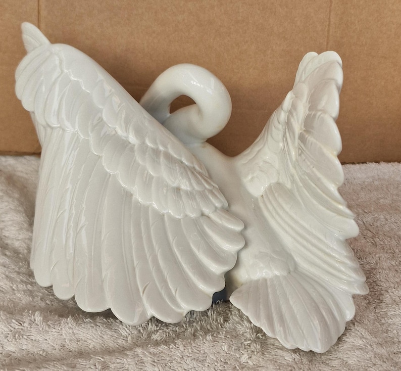 May include: A white ceramic swan figurine with outstretched wings. The swan's wings are detailed with individual feather carvings. The swan's neck is curved, and the head is turned back. The figurine is a decorative item.